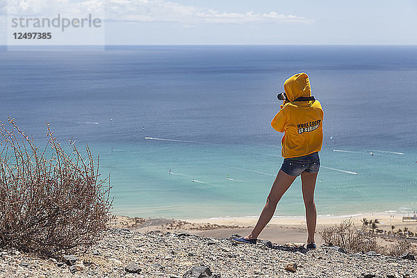 Mädchen  das von einer hohen Klippe aus einen Strand fotografiert. Kapuzenpulli/Pullover