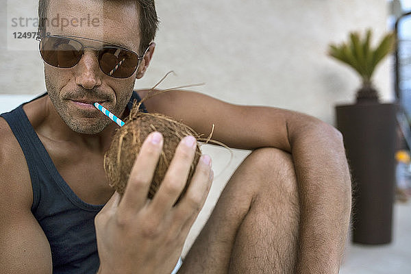 Nahaufnahme eines Mannes  der eine Sonnenbrille trägt und eine Kokosnuss aus einem Strohhalm trinkt