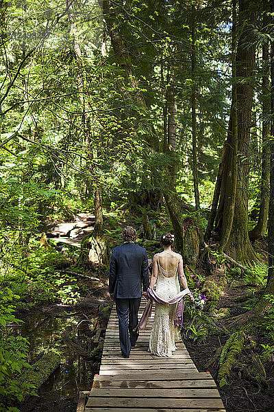 Ein junges Ehepaar hält sich an den Händen  während es über eine Uferpromenade im Wald geht.