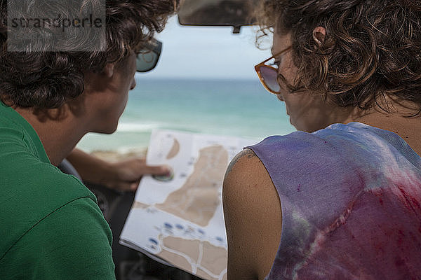 Ein junges Paar (25-35 Jahre alt) sitzt in seinem Auto und schaut auf eine Landkarte mit dem Meer im Hintergrund.