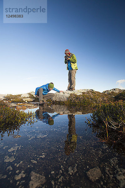 Eine junge Frau erkundet das Mikroklima eines Bergsees  während ihr Partner ein Foto von ihr schießt.