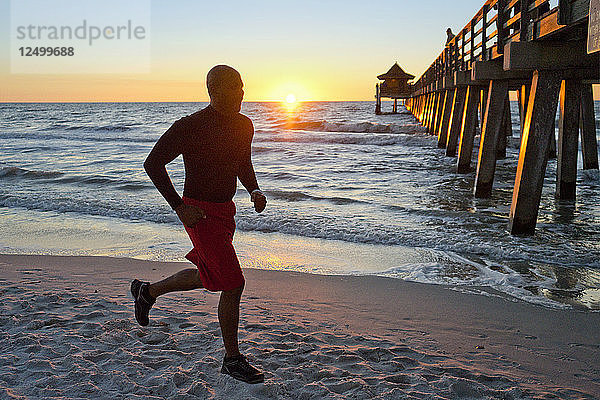Mann läuft am Strand bei Sonnenuntergang mit einem Pier im Hintergrund