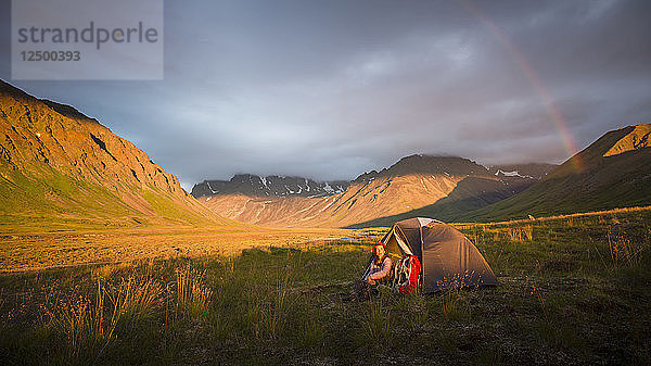 Eine Frau sitzt bei ihrem Zelt  als ein Regenbogen über dem Camp im Lake Clark National Park and Preserve aufzieht