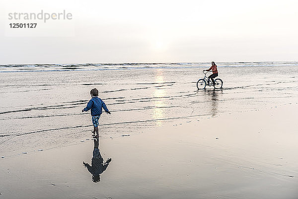 Eine Frau fährt mit dem Fahrrad  während ihr kleiner Junge am Strand spielt.