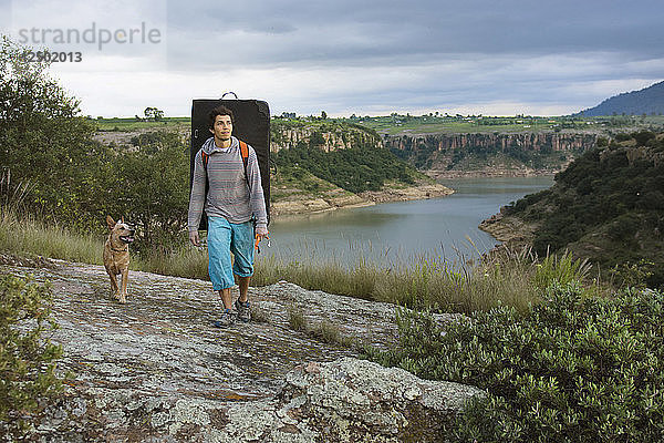 Ein männlicher Bergsteiger  der ein Crash Pad trägt  erkundet mit seinem Hund die Umgebung eines Sees