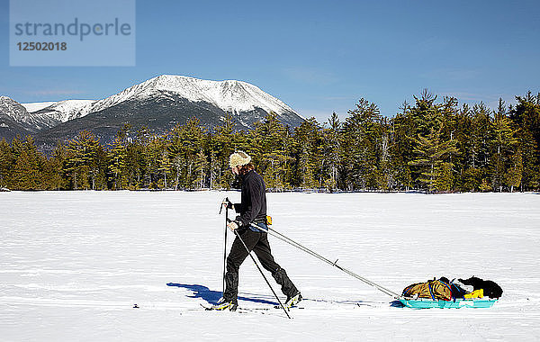 Ein Mann fährt Langlaufski über einen zugefrorenen See  während er einen Pulk-Schlitten zieht. Im Hintergrund ist der Mount Katahdin zu sehen.