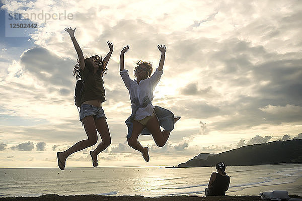 Junge Frauen springen mit erhobenen Armen an der Küste