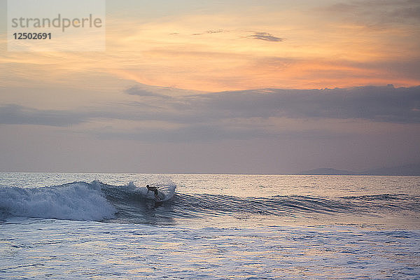 Ein orangefarbener Himmel begleitet einen männlichen Surfer  der auf einer kleinen Welle zum Strand reitet.