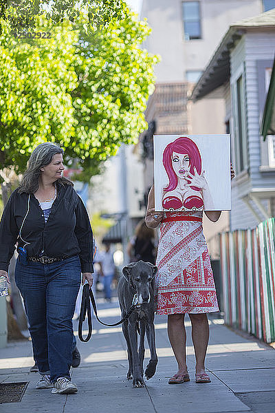 Eine Frau hält in San Diegos Little Italy ein kaputtes Gemälde vor sich  während eine Passantin und ihr Hund neugierig zuschauen.