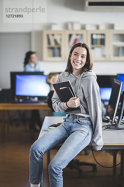 Cheerful high school female student sitting with books on desk in classroom