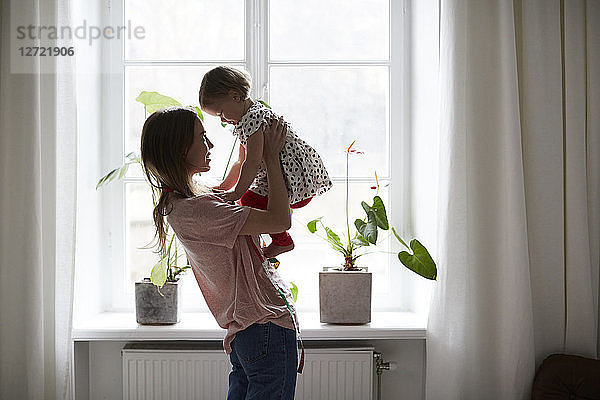 Side view of fashion designer carrying daughter while standing by window at home