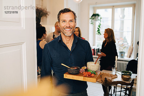 Portrait of smiling mature man holding food in serving tray while standing against friends at party