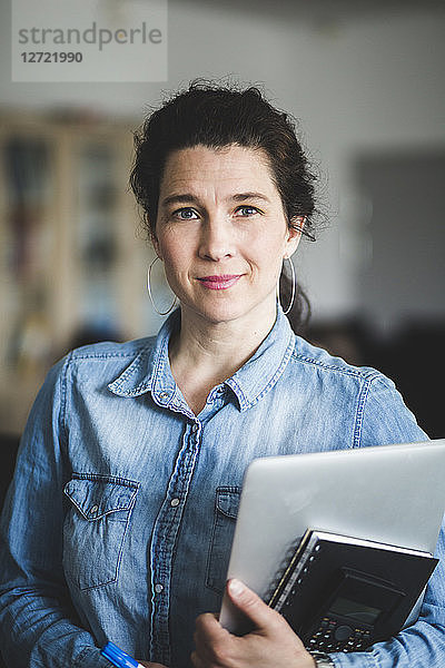 Portrait of confident mature female teacher holding diary and laptop while standing at computer lab