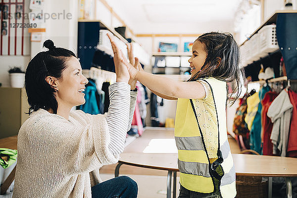 Side view of happy teacher and girl giving high-five in cloakroom at kindergarten