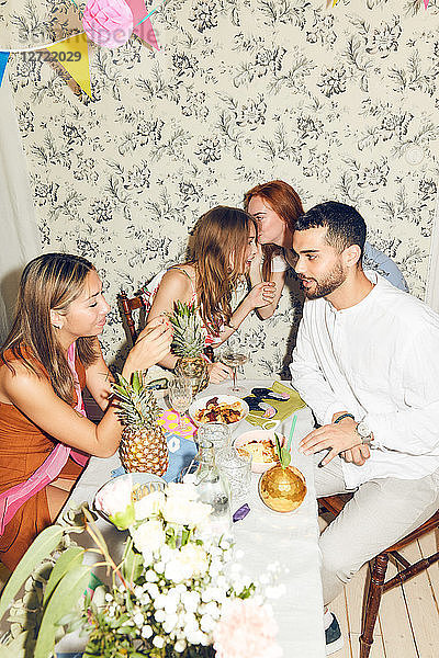 Young female friends whispering while man and woman sitting at dining table during party in apartment