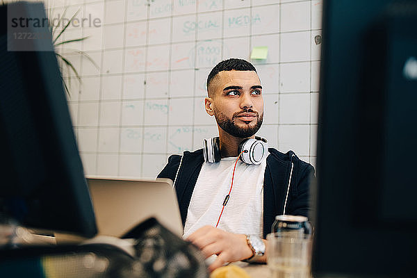 Young male computer hacker sitting with laptop while looking away at creative office