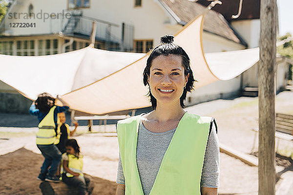 Portrait of confident female teacher standing in reflective clothing against students playing at background