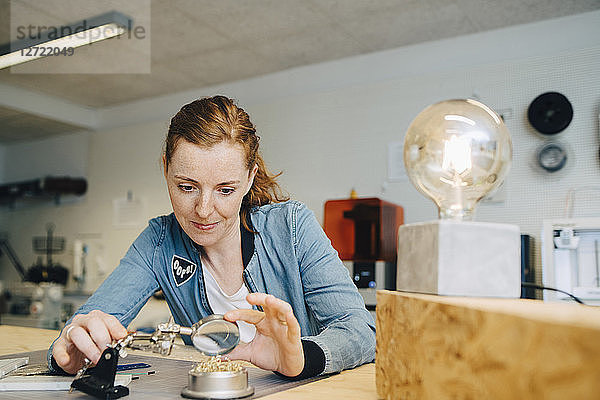 Confident redhead female engineer looking through magnifying glass at workbench in creative office