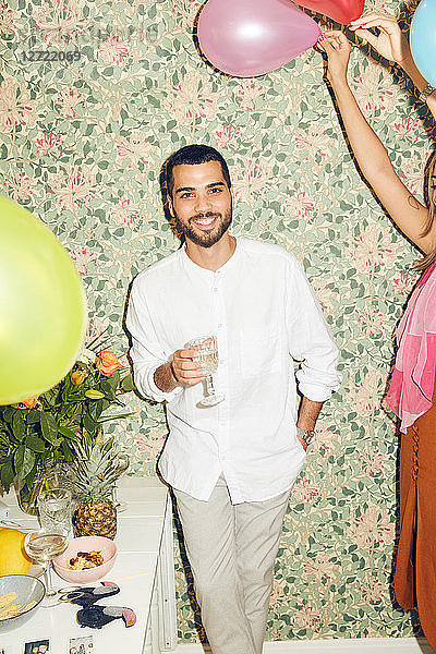 Portrait of smiling young man holding drink while standing by woman with balloons against wallpaper at home during dinne