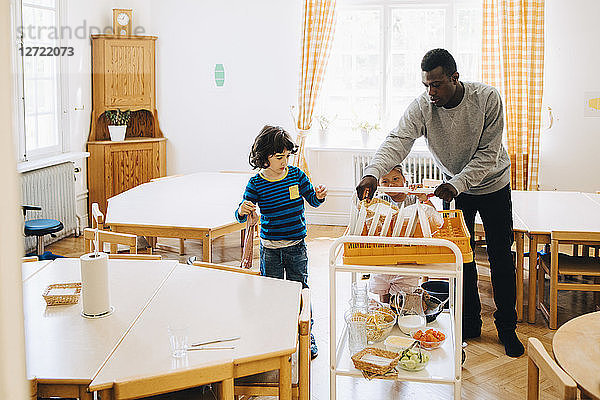 Male teacher assisting students pushing cart amidst tables in classroom at child care