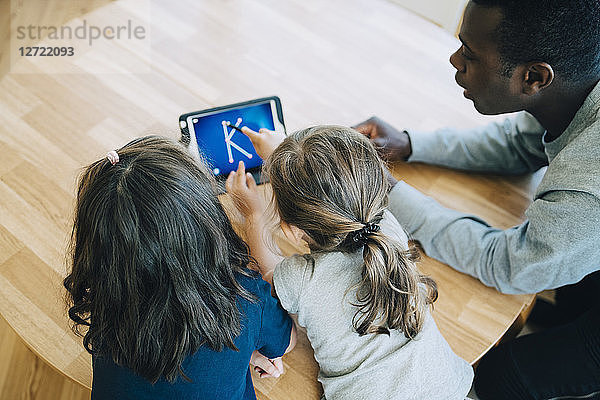 High angle view of girl touching letter K on digital tablet amidst friend and teacher at table in classroom