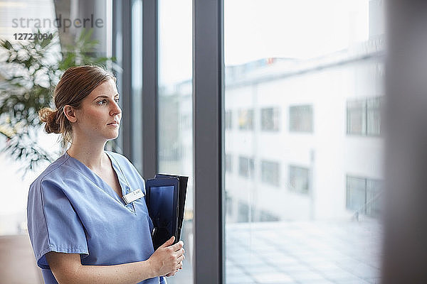 Thoughtful female nurse with digital tablet looking through window while standing in corridor at hospital
