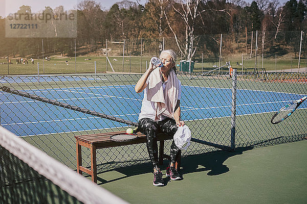 Tired senior woman drinking water while sitting on bench at tennis court