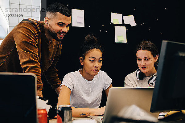 Colleagues looking at businesswoman using laptop against bulletin board at creative office