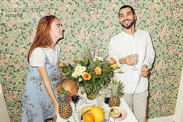 Smiling young friends standing by table against wallpaper during dinner party at home