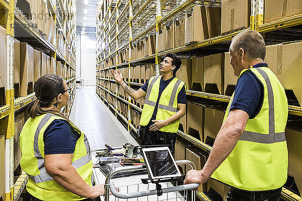 Young worker discussing with coworkers with cart while standing on aisle amidst racks at distribution warehouse