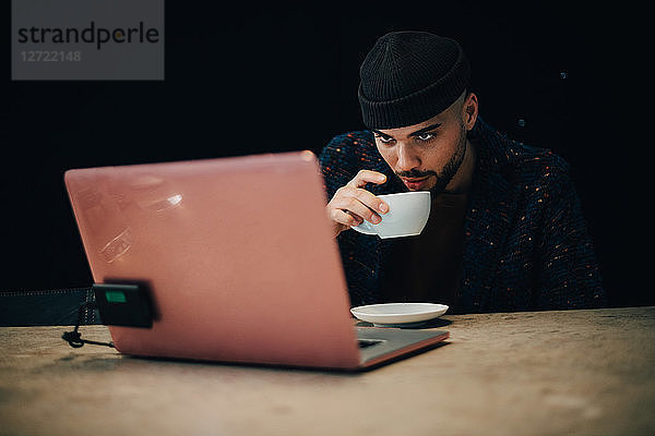 Confident young male hacker drinking coffee while looking at laptop on desk in office