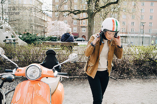 Young businesswoman removing helmet while walking by motor scooter on roadside in city