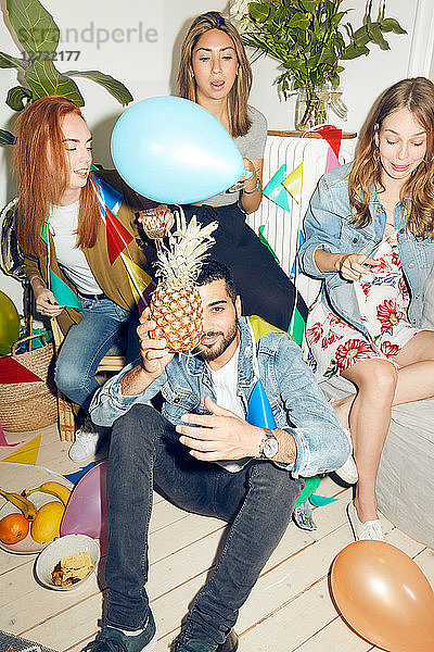 Portrait of young man holding pineapple while sitting against female friends enjoying party at home