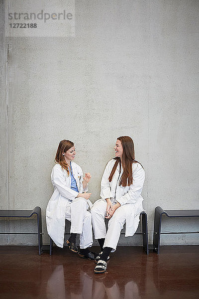 Full length of smiling female doctors talking while sitting against wall at hospital
