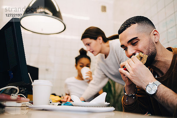 Young male hacker eating sandwich while female colleagues working during meeting at small office