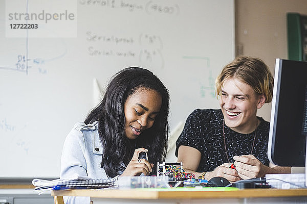 Smiling female student preparing science project with young male friend on desk in classroom at high school