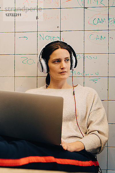 Young female computer hacker listening through headphones while sitting with laptop against tile wall at creative office