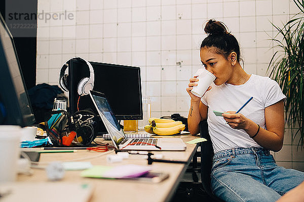 Young businesswoman drinking coffee while sitting at desk in small creative office