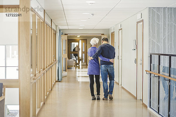 Rear view of senior woman walking with son in corridor at nursing home