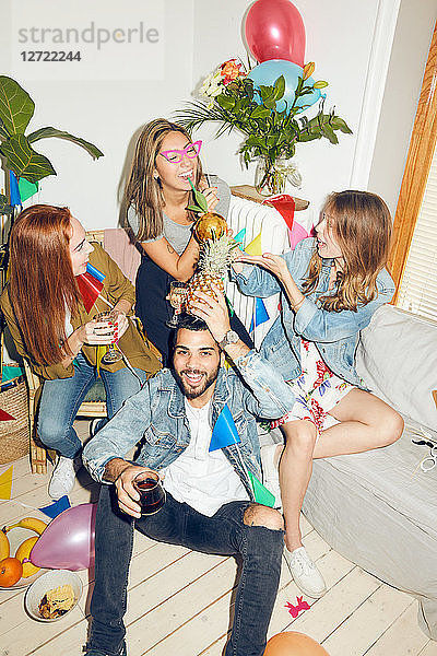 Portrait of smiling young man holding pineapple while sitting against female friends enjoying party at home