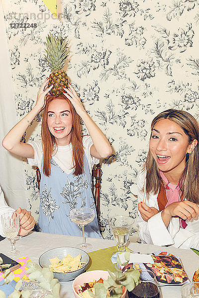 Playful young woman holding pineapple on head while sitting with friends at dining table during party in apartment