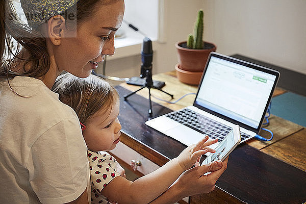 Podcaster showing mobile phone to daughter while using laptop at home