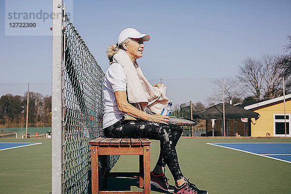 Side view of tired senior woman sitting on bench against sky at tennis court