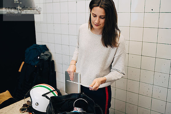 Young busineswoman removing laptop from bag while standing against wall at small office