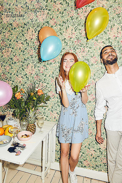 Young man and woman playing with balloons while standing against wallpaper at home during dinner party