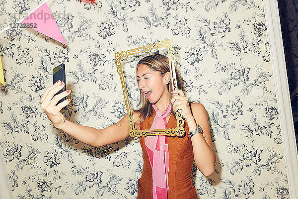 Cheerful young woman taking selfie through picture frame while winking at smart phone against wallpaper during party in