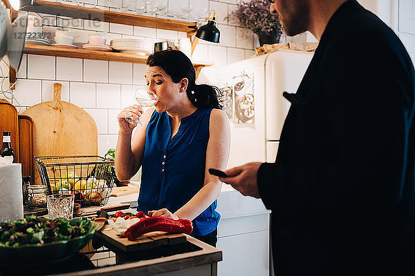 Male friend looking at mature woman drinking wine while standing in kitchen