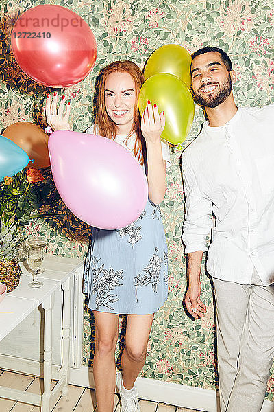 Portrait of smiling young woman playing with balloons while standing by man against wallpaper at home during dinner part