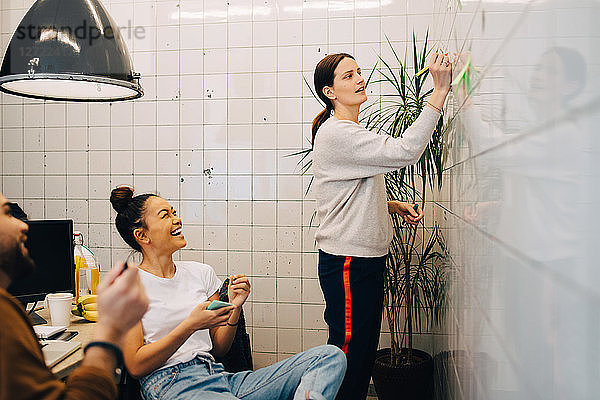 Smiling colleagues looking at young businesswoman writing strategy on tile wall in creative office