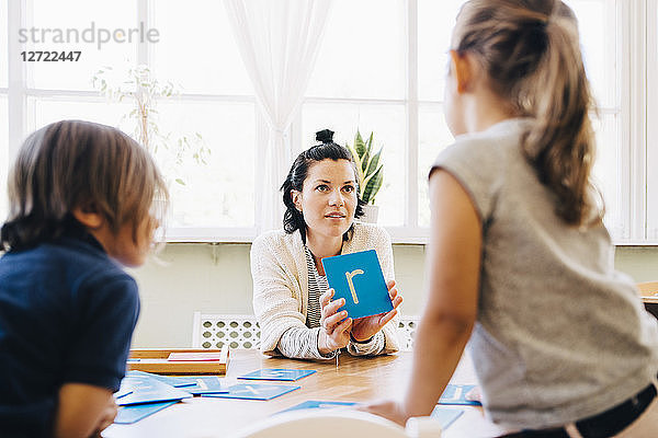 Confident female teacher showing letter R to students at table in classroom
